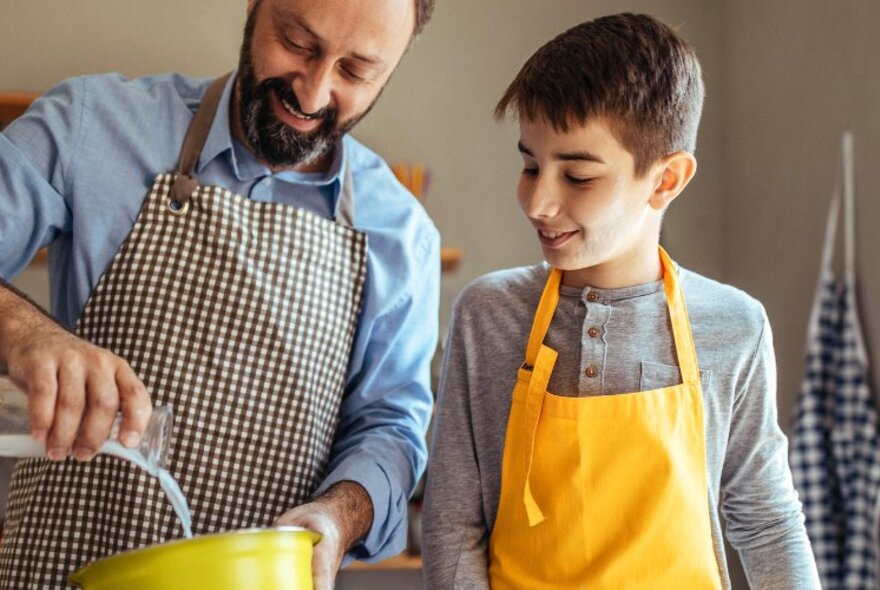 A father and his young teenage son, both wearing aprons, cooking in a workshop.