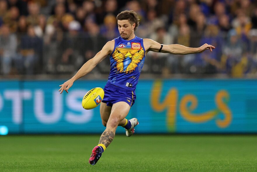 A West Coast Eagles AFL player running on the field, about to kick a football that hovers in front of him, a blurred stadium crowd behind him.