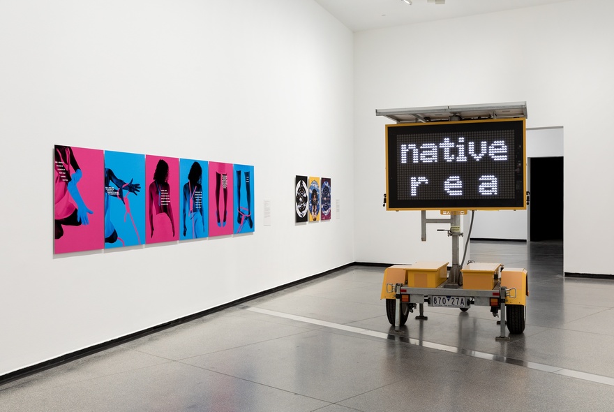 A gallery with white walls and grey floors, with a series of bot pink and blue artworks on the left wall, a digital roadside sign displaying the words, 'native r e a'.