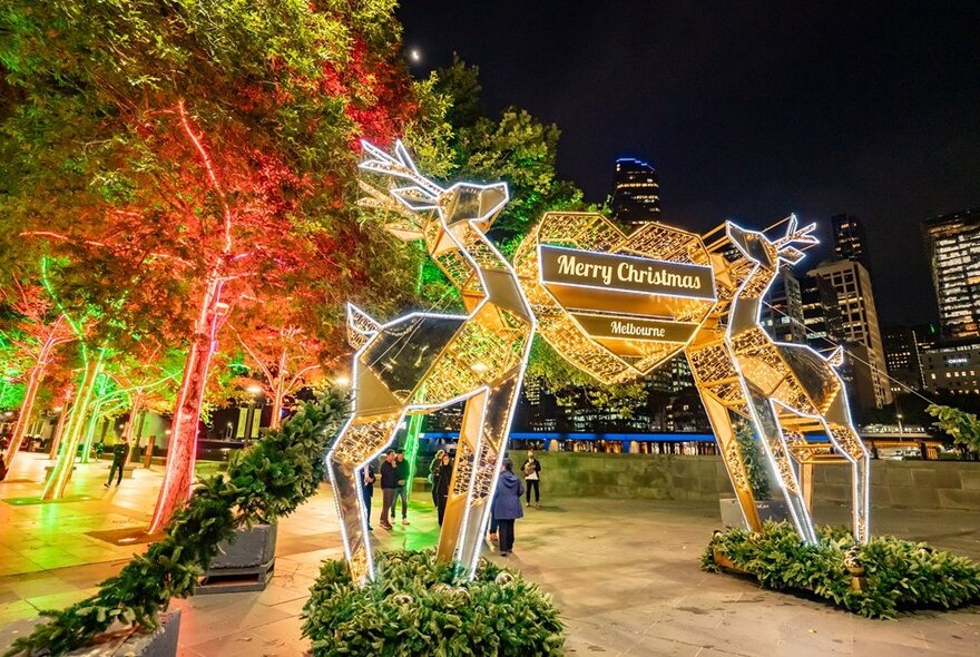 A glowing arch of two reindeer joined by a gold heart with 'Merry Christmas Melbourne' text, surrounded by green foliage.
