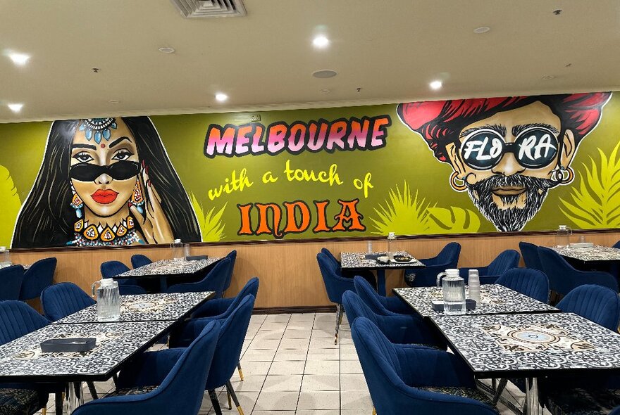 The interior of an Indian Restaurant with a painted mural on the back wall, blue velvet chairs and mosaic tables.