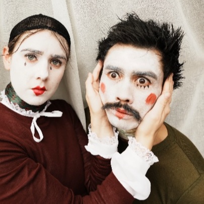 Man and woman wearing theatrical white makeup with lipstick and dark eye details, woman holding the man's face in her hands.