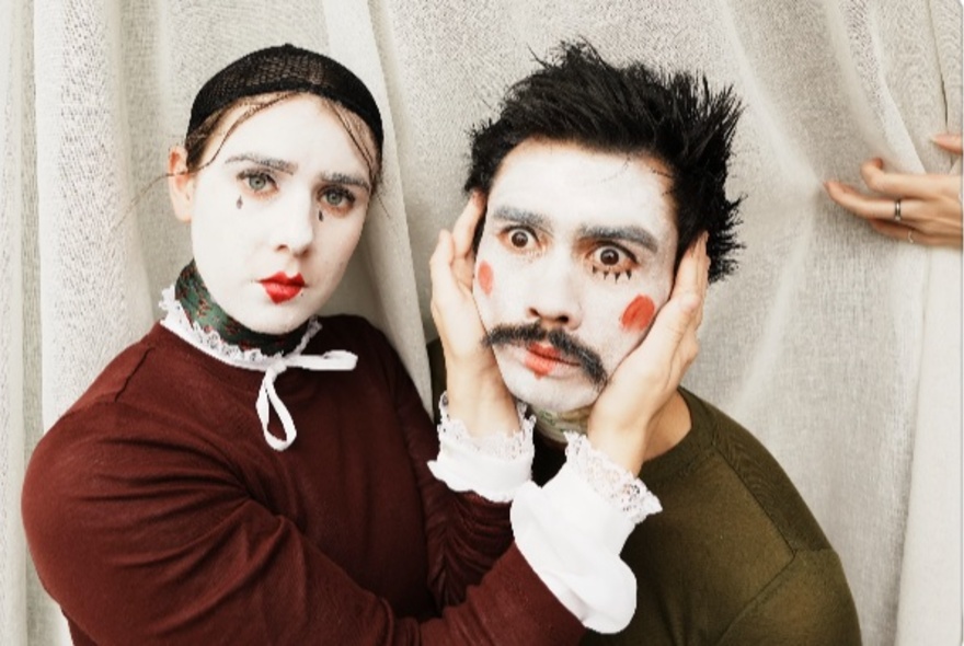Man and woman wearing theatrical white makeup with lipstick and dark eye details, woman holding the man's face in her hands.
