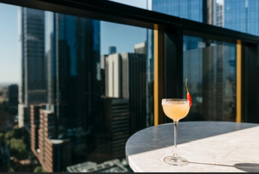 A single long-stemmed cocktail glass on a round table, with city buildings behind.