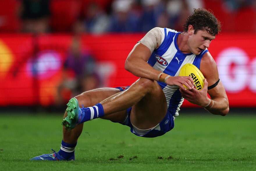 A North Melbourne AFL player holding a yellow football as he falls to the ground during a game.