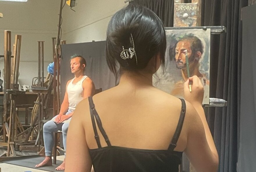 A clothed life model seated on a stage in a studio setting, while in the foreground a young artist uses a brush to paint this scene onto a canvas.