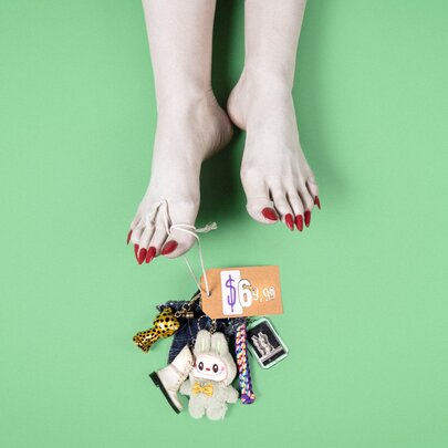 A pair of bare feet with pointed red toenails positioned on a solid green background, an ID tag hanging from the big toe, directly above a messy cluster of keychains, a plush toy, and an orange tag attached to a ring.