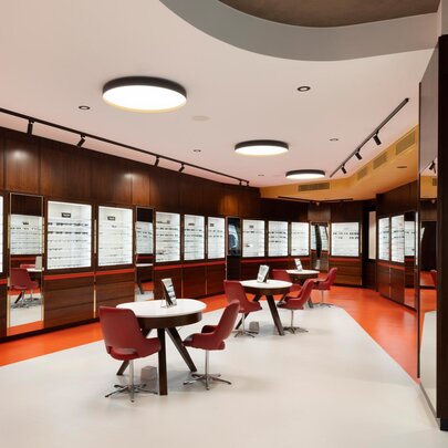 Opticians retail store with walls lined with shelves of spectacles and central chairs and tables.