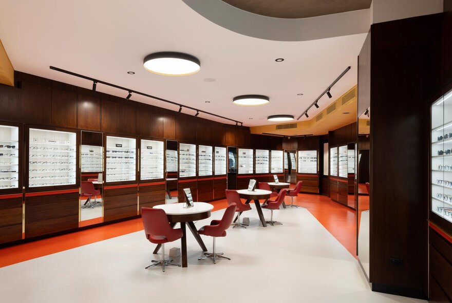 Opticians retail store with walls lined with shelves of spectacles and central chairs and tables.