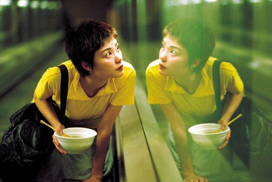 An Asian woman wearing a yellow t-shirt and holding a bowl of noodles, looking at her reflection in a glass window.