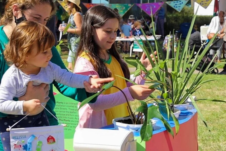 Two children and their adult carer interacting with green plants and touching their leaves, at the RESET Festival at Fed Square.