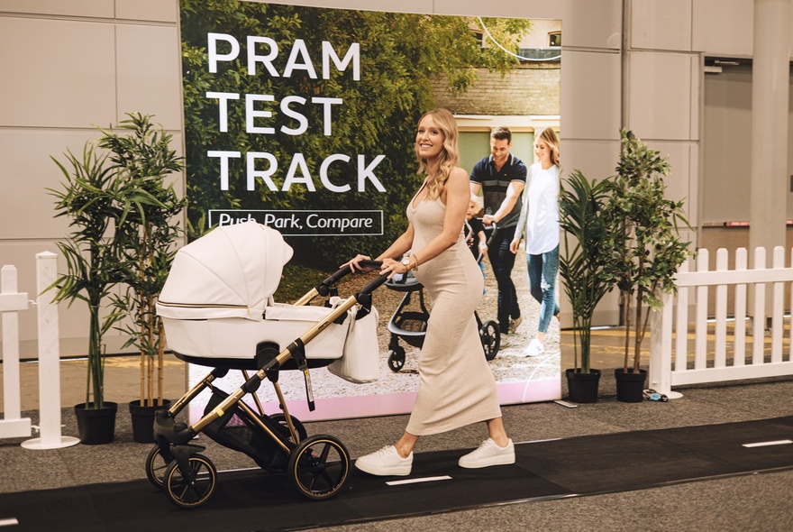 A pregnant woman pushing a pram on a track tin front of a sign that reads PRAM TEST TRACK at The Pregnancy Babies & Children's Expo.