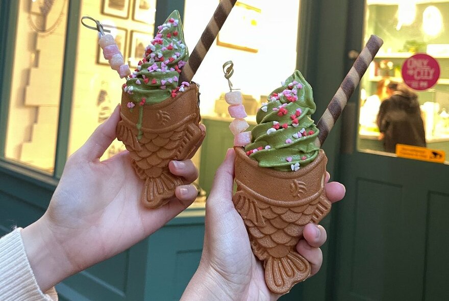 Two hands holding up waffles shaped like fish topped with soft-serve matcha ice-cream, sprinkles and a biscuit stick, in front of a shop window. 