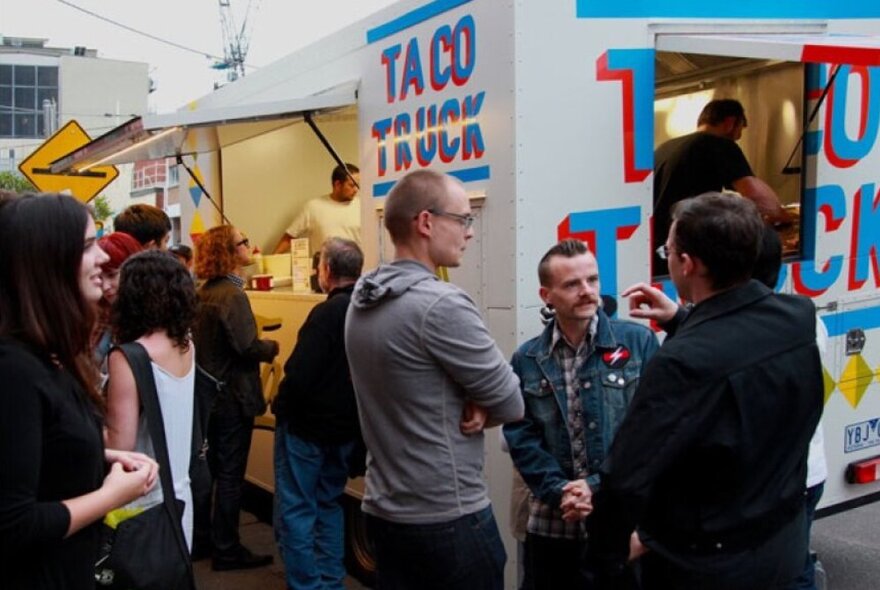 A group of people talking and waiting for food in front of a taco food truck.