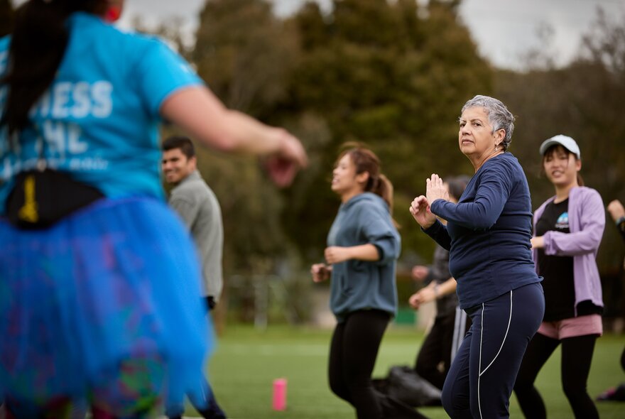An outdoor group fitness class in a park with several participants of different ages exercising on the grass.
