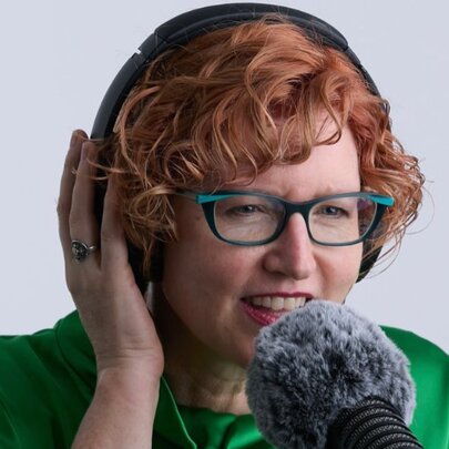 A woman with short, curly reddish-brown hair wearing glasses and over-ear headphones, speaks into a fuzzy microphone in the foreground.