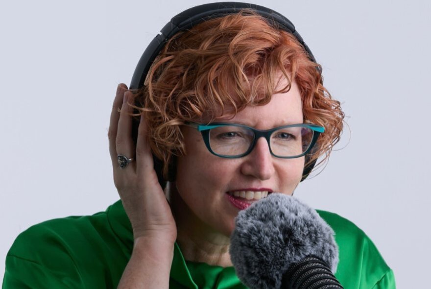 A woman with short, curly reddish-brown hair wearing glasses and over-ear headphones, speaks into a fuzzy microphone in the foreground.