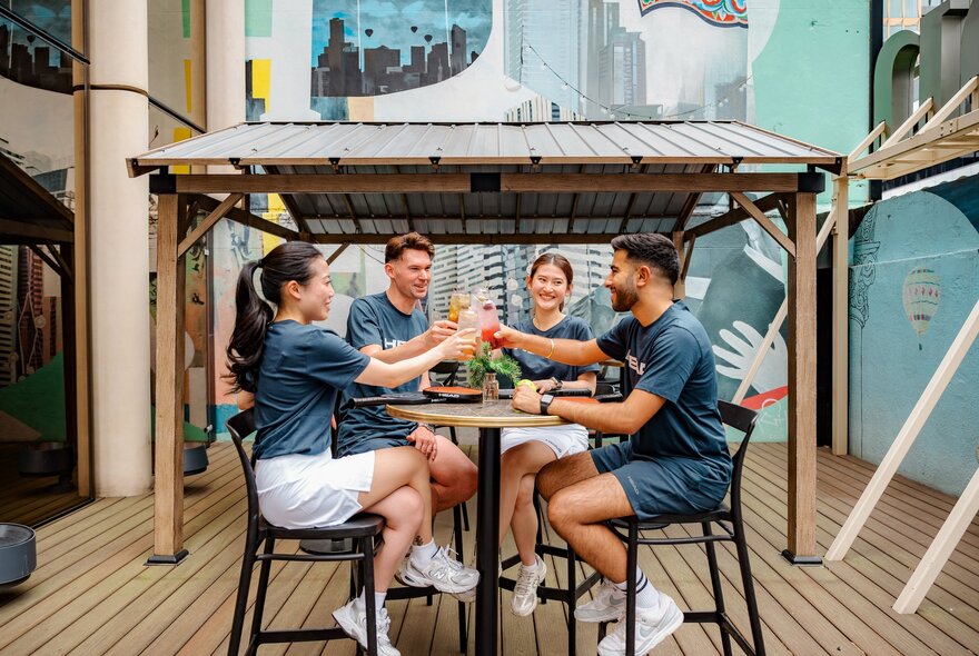 Four people in matching blue shirts sitting at a high outdoor table, toasting with drinks and smiling.