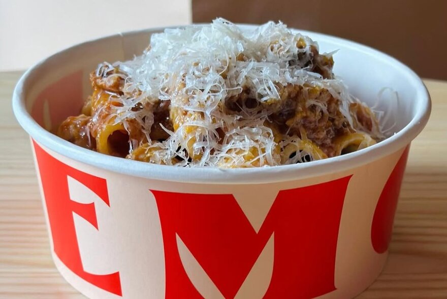 A cardboard takeaway bowl filled with a traditional Italian bolognese with penne, sprinkled with parmesan cheese.