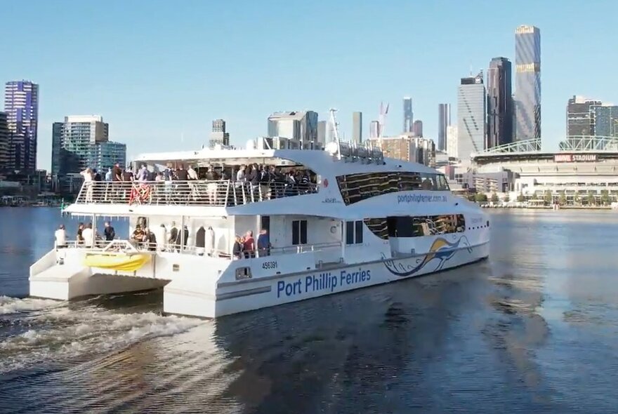 A large ferry with people on the top deck cruising on the Yarra River with the Melbourne skyline in the background. 