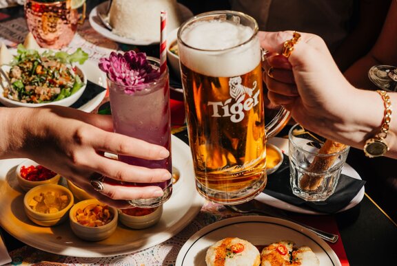 People clinking glasses of Tiger beer and a cocktail over a table spread with Thai food. 