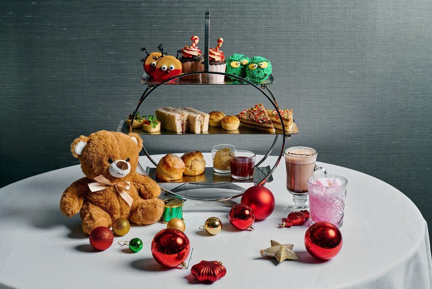 A children's themed three-tiered platter of high tea treats, alongside a teddy bear, drinks and Christmas baubles.