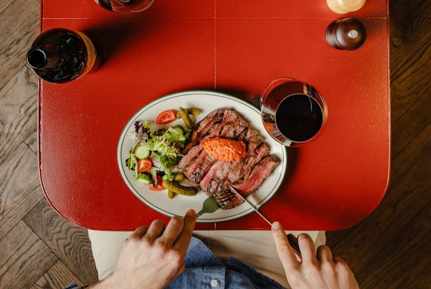 Hands holding cutlery cutting into a piece of meat on a plate with salad and glass of wine.