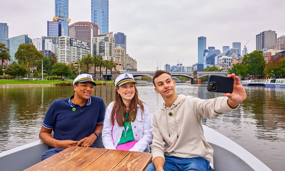 Three people taking a selfie on a small boat on the Yarra River with the city in the background.