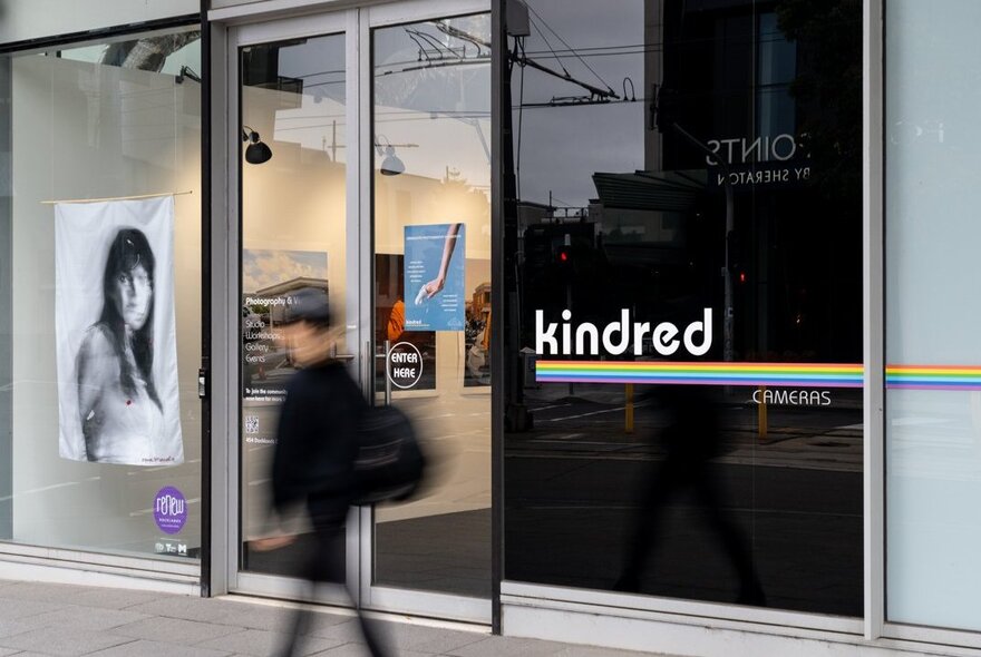 The exterior of Kindred Gallery with an artwork on display in the large window, and a pedestrian walking past.