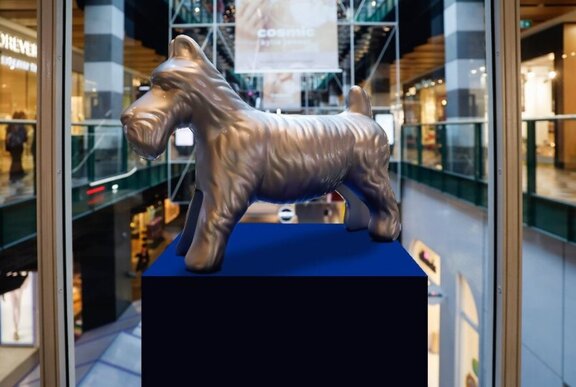 Silver dog Monopoly game piece on a blue plinth inside a glass display case in a shopping mall.