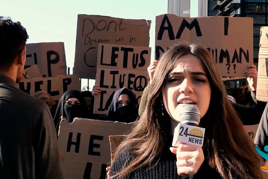 Still from a film depicting a journalist talking into a microphone while reporting from a street protest, people with placards standing behind her.