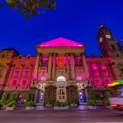 The facade of Melbourne Town Hall illuminated in vibrant pink and purple at dusk, with a blurred tram going past.
