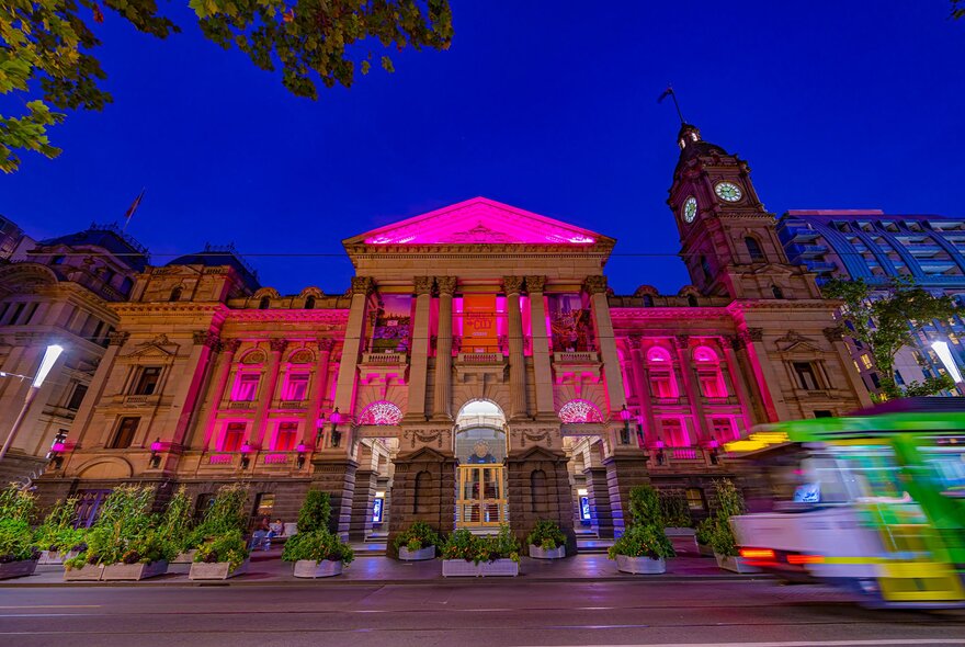 The facade of Melbourne Town Hall illuminated in vibrant pink and purple at dusk, with a blurred tram going past.