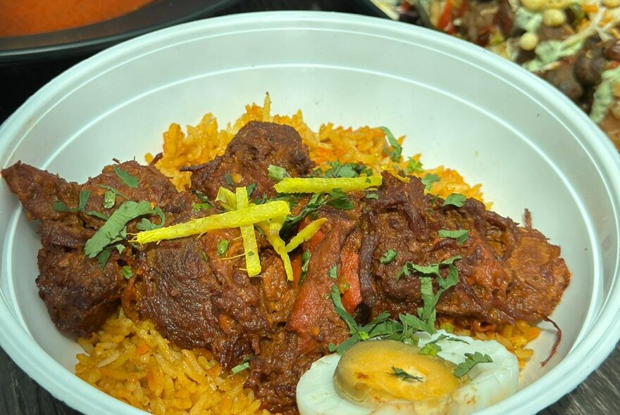 An Indian beef dish in a takeaway bowl, a fried egg on the side.