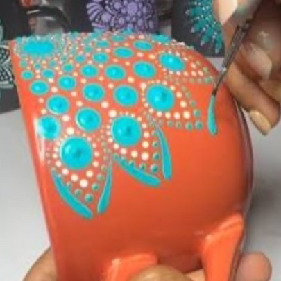 A person using a small brush to create a dot pattern design with turquoise and white acrylic paint on an orange ceramic mug, as part of a creative workshop.