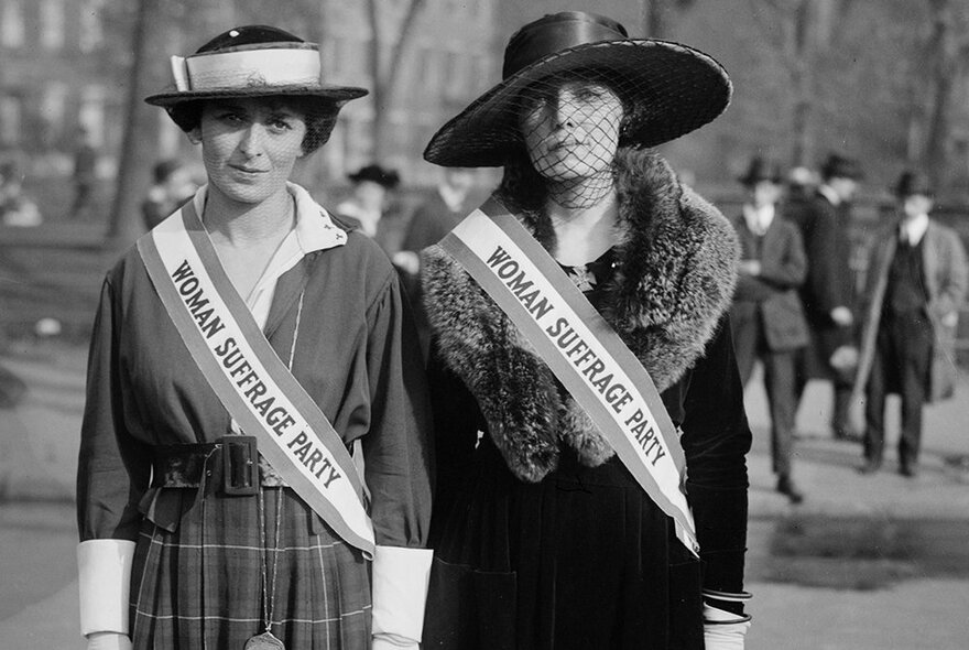 Historic photograph of two suffragettes wearing sashes with men in top hats behind them looking on.