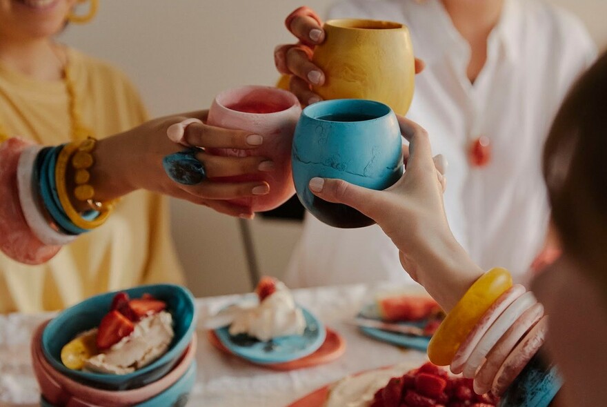 Three people wearing designer jewellery and holding brightly-coloured mugs. 