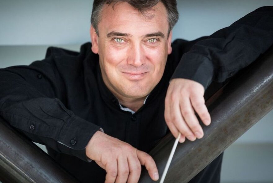 Orchestra conductor Jaime Martín smiling and holding a conducting baton in his hand, leaning forward and resting his arms on a wooden beam.