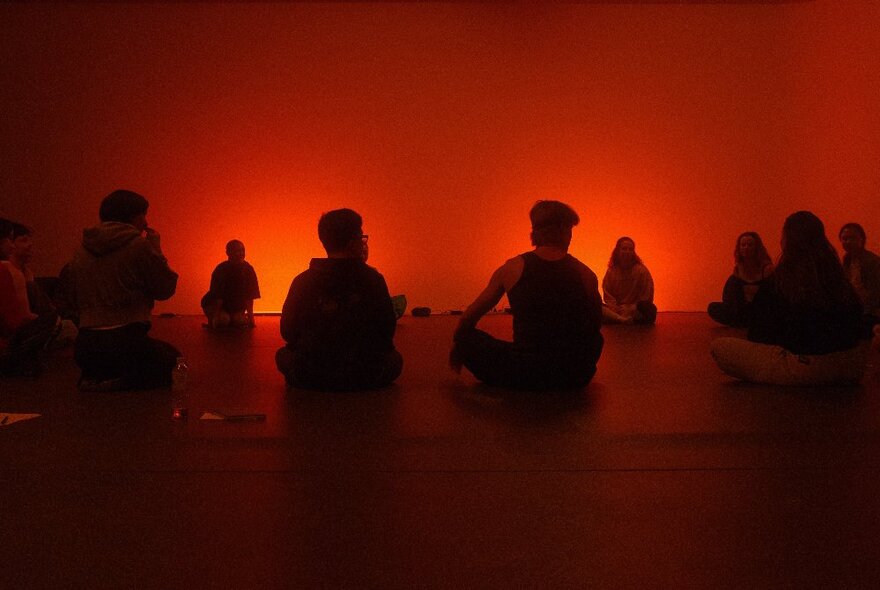 A group of people sitting cross legged on the floor of a room that is bathed in orange-red light.