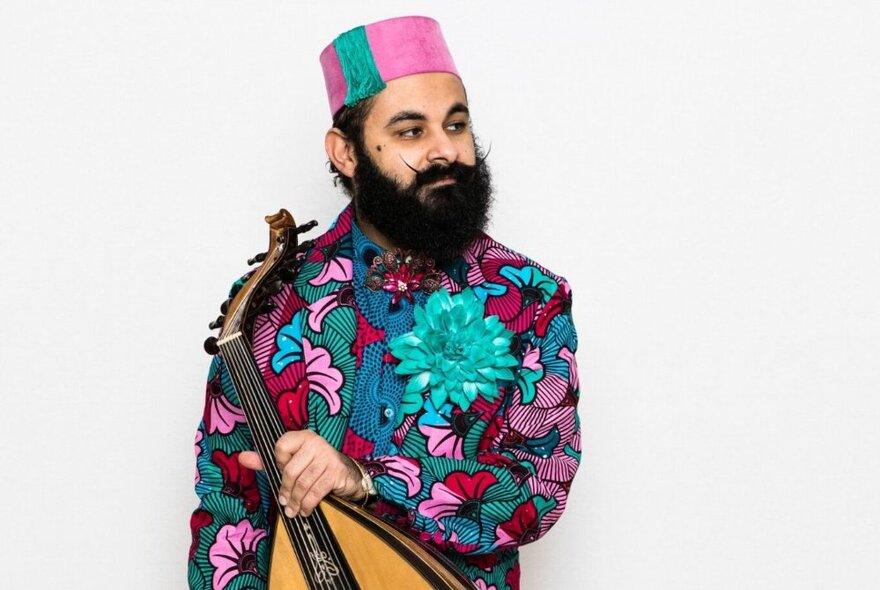 Joseph Tawadros, wearing a brightly patterned shirt and fez, holding his oud and looking off to the side of the frame, standing against a white background.