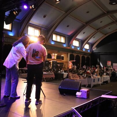 Two people standing on a stage that looks out to a large hall, people seated at tables and chairs in the space.