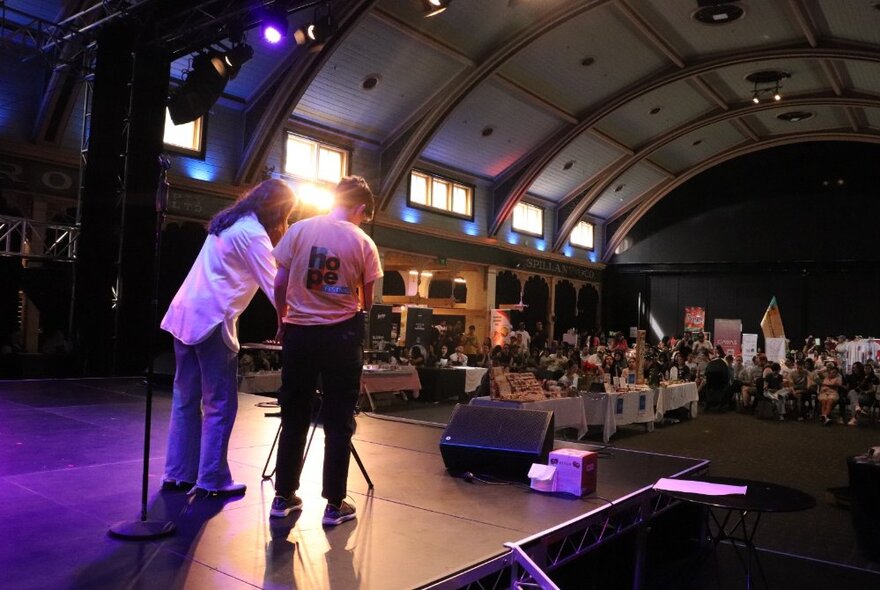 Two people standing on a stage that looks out to a large hall, people seated at tables and chairs in the space.