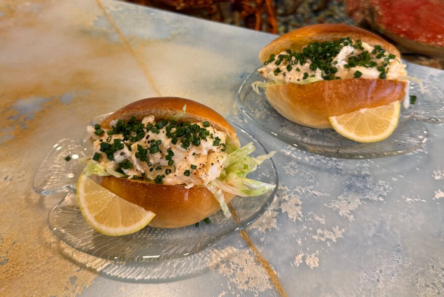 Two lobster rolls presented on fish-shaped glass plates on a marble table.