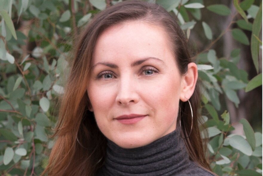 A headshot of a woman in a polo neck jumper and large hoop earrings in front of a leafy bush.