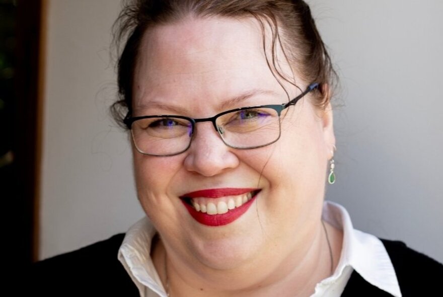Smiling face of a woman wearing glasses, their hair pulled back from their face, wearing bright red lipstick.