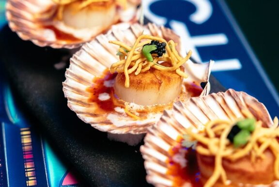 Close up of three seared scallops in their shells with a garnish of dry fried noodles.