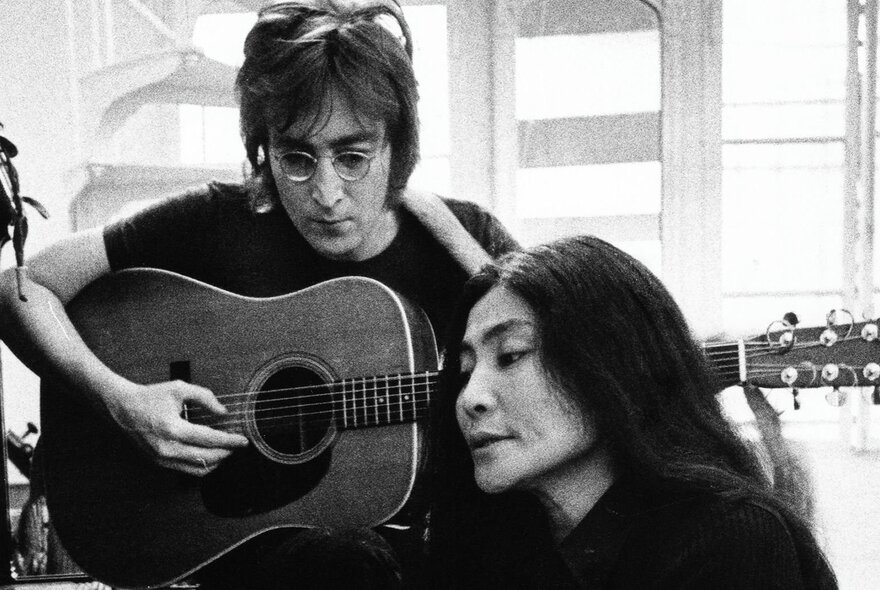 A black and white image of John Lennon playing guitar behind Yoko Ono