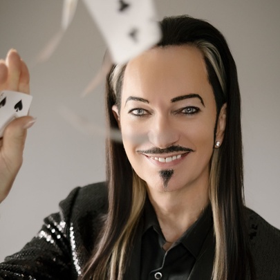 A long-haired man with a waxy appearance, moustache and pointy little goatee smiles has he flicks playing cards, one-handed. 