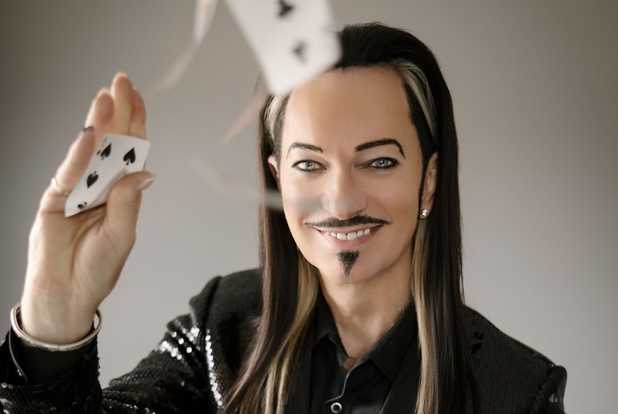 A long-haired man with a waxy appearance, moustache and pointy little goatee smiles has he flicks playing cards, one-handed. 