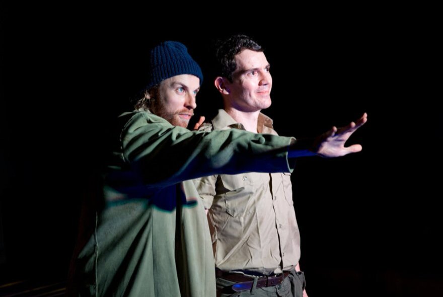 Two actors spot lit on a stage, both looking out toward the audience, one with his arm stretched out in front of him.